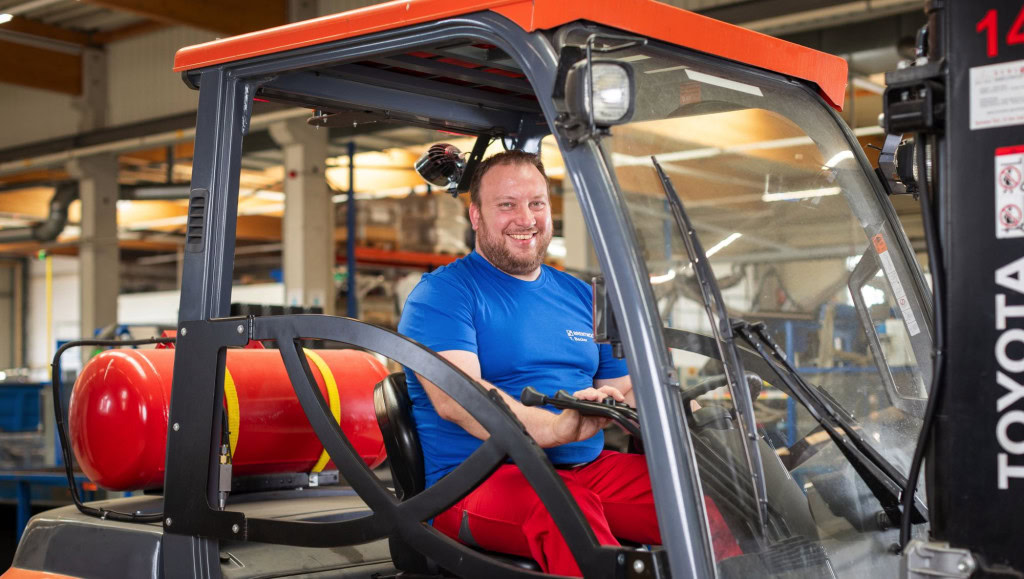 A man with a beard, wearing a blue Brentwood Industries shirt and red pants, smiles while sitting in the driver's seat of a forklift inside a warehouse in Wettringen, Germany.