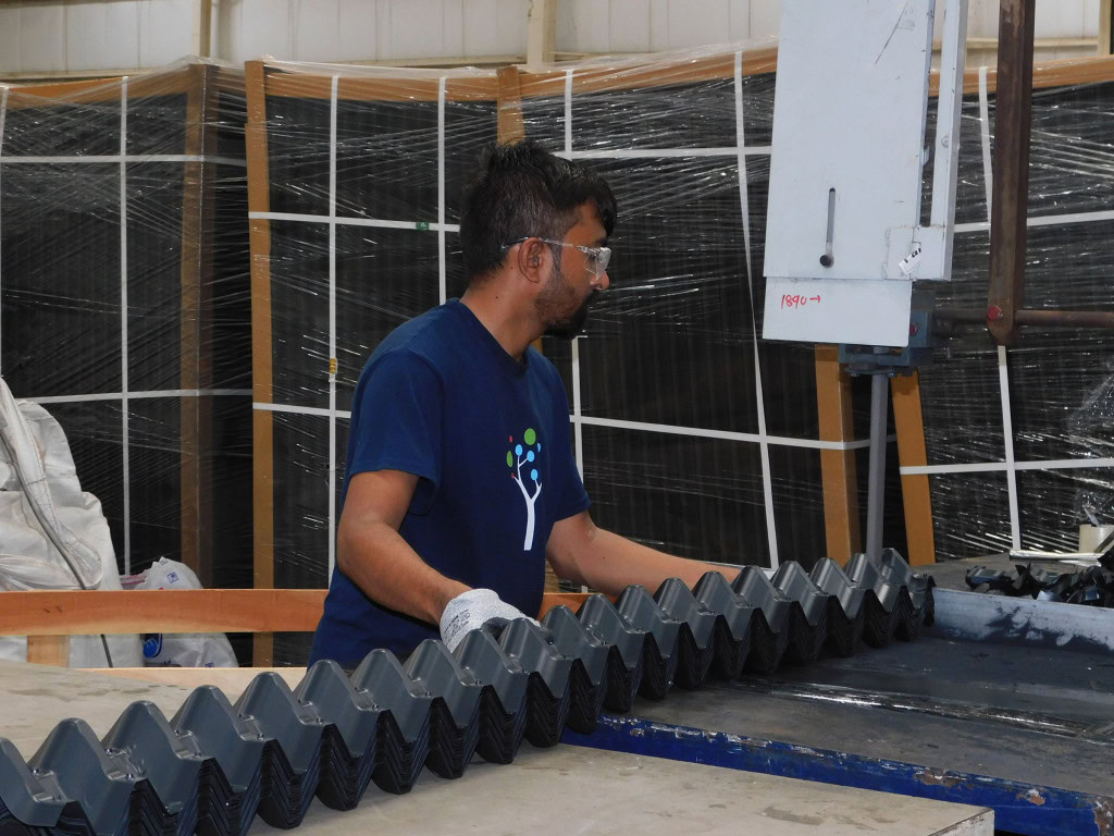 A man wearing safety glasses and gloves is working at a machine in a factory. He is handling a row of identical, angular parts on a worktable.