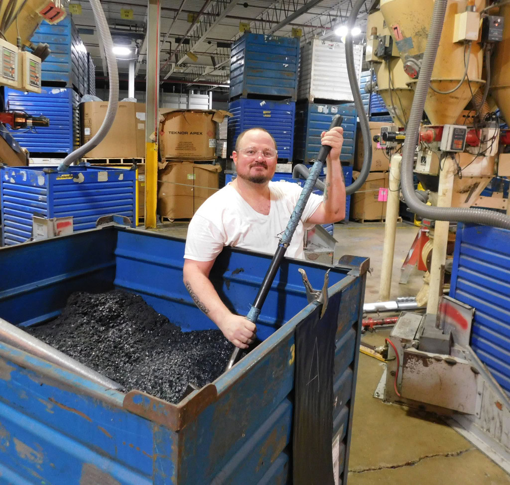 A man in a white T-shirt stands in a factory beside a large blue bin filled with dark material, holding a tool. About him, several other blue bins and pieces of industrial equipment are scattered.