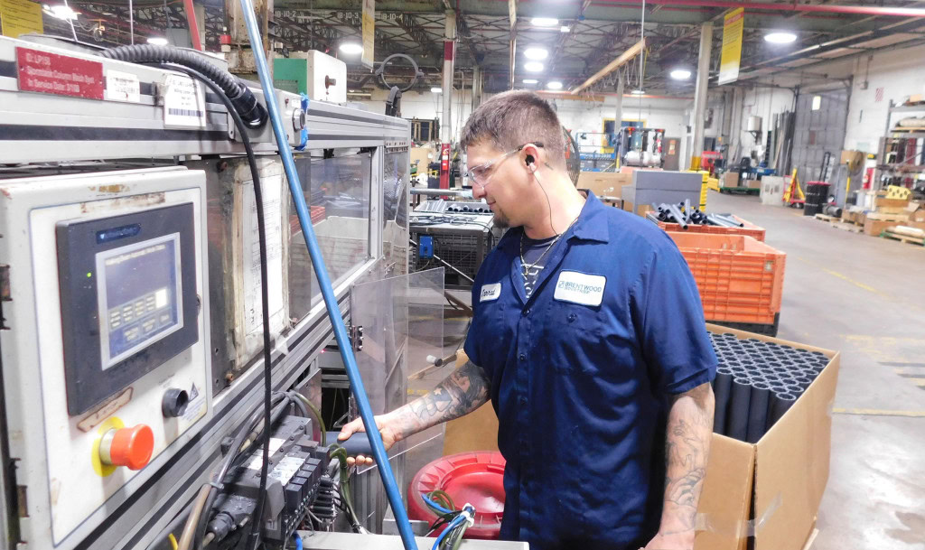 A man wearing safety glasses and ear protection operates machinery inside an industrial setting. He is dressed in a dark blue uniform shirt and surrounded by various equipment and supplies.