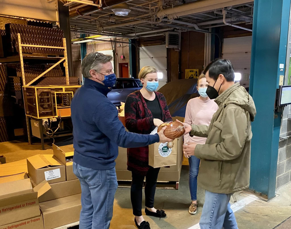 Four people are gathered in a warehouse, with two handing over a wrapped turkey and food items to the others. Everyone is wearing face masks. Boxes are stacked in the background, reflecting a community culture of giving at this food distribution event.