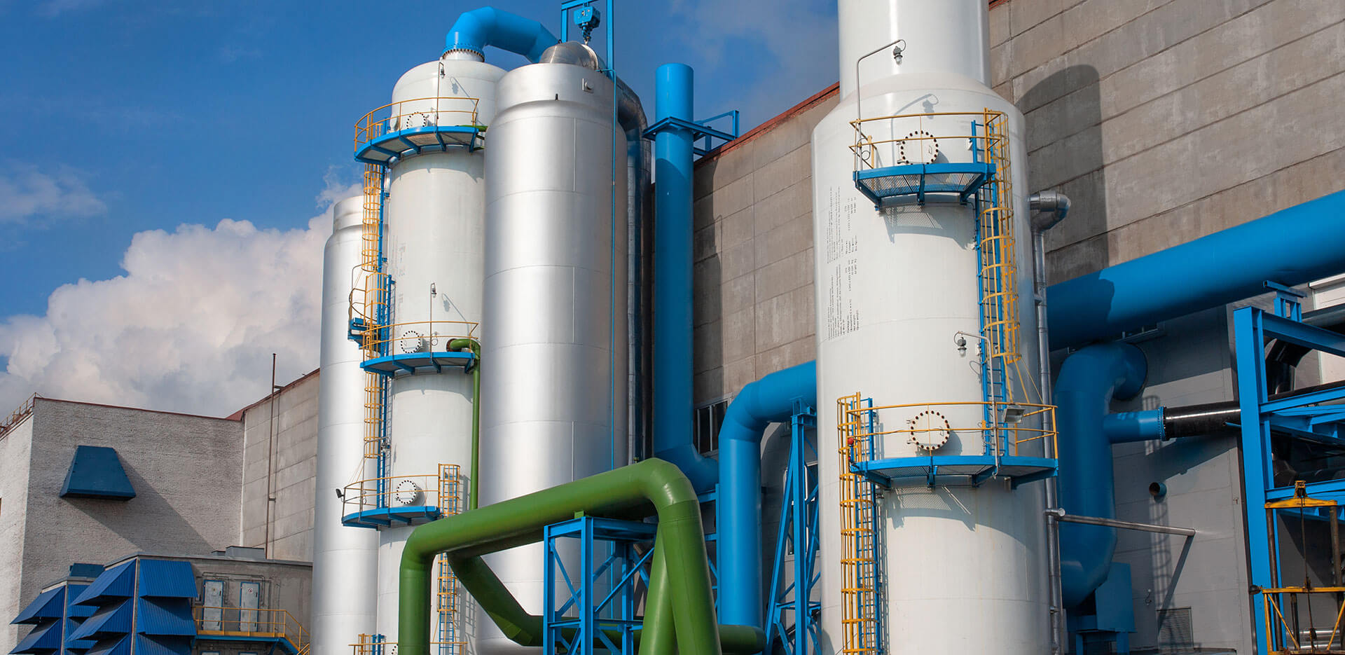 An industrial facility with tall cylindrical metal tanks, fitted with blue and green pipes against a cloudy sky backdrop. The tanks and pipes feature yellow ladders and railings.