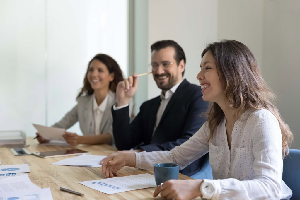 Three colleagues sitting at a conference table during a meeting, smiling and engaged. They have papers, a pen, and a coffee mug in front of them.