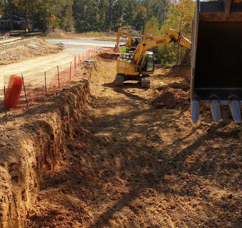 A large excavator digs a deep trench at a construction site, playing a crucial role in stormwater management. Another excavator is visible in the background. The area is bordered by orange safety mesh fencing, and trees are visible in the distance.