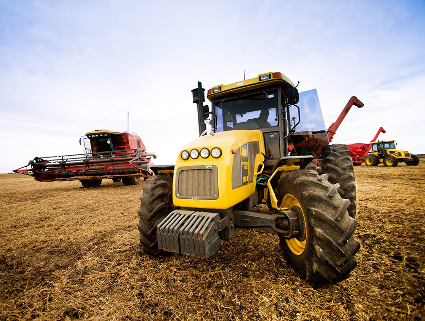 A yellow tractor is prominently displayed in the foreground on a wide, expansive field covered with hay or crop residue. In the background, there is a red combine harvester and another yellow vehicle.