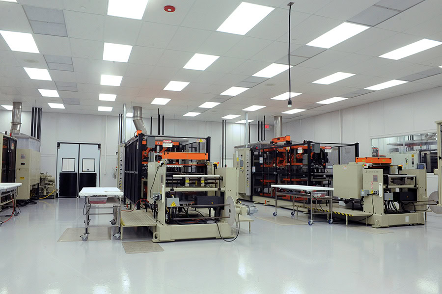 Large industrial room with bright overhead lighting, several large industrial machines, and conveyor belts. The machines are predominantly beige with orange and black components.