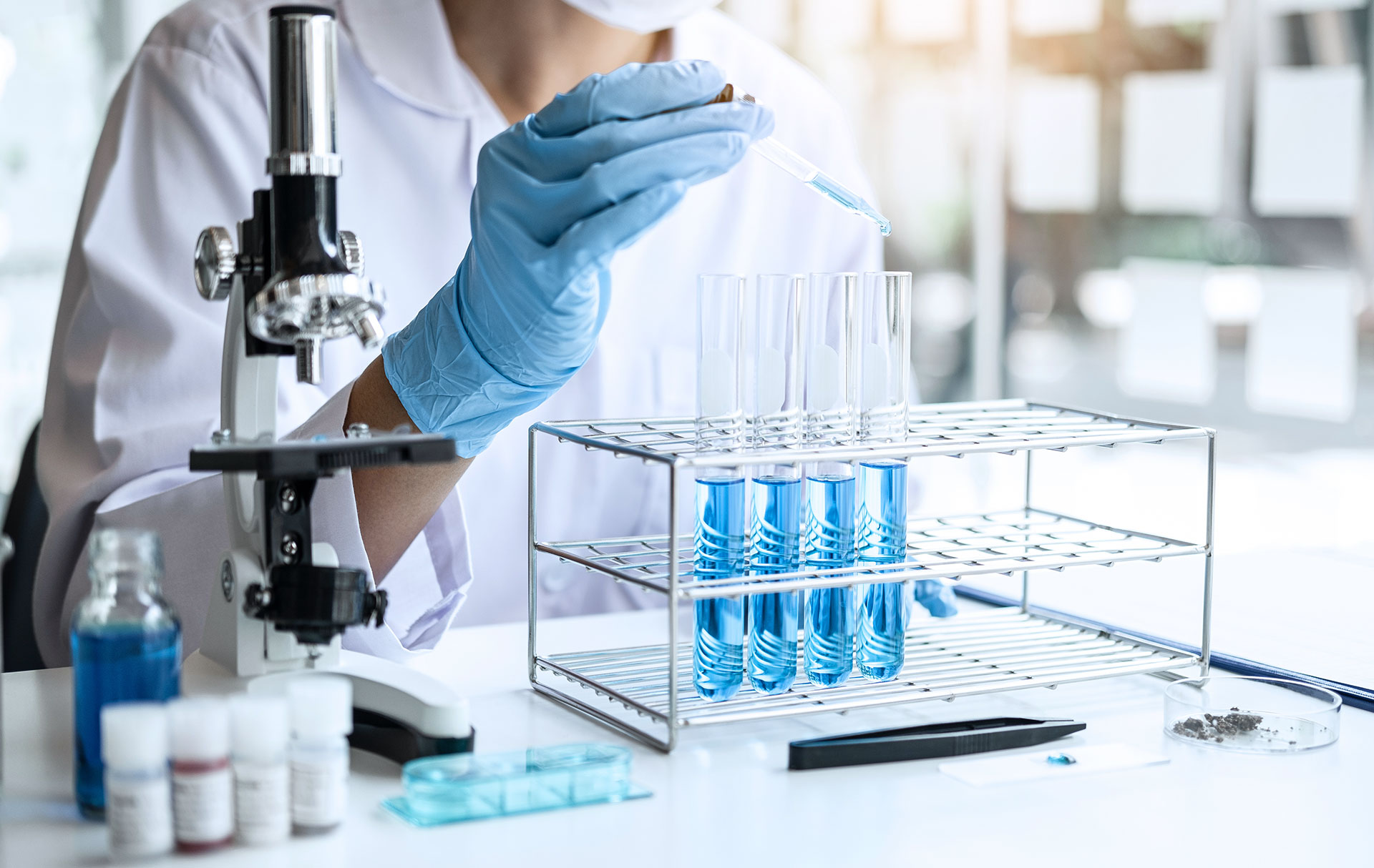 A scientist wearing a lab coat and blue gloves uses a dropper to add liquid to one of several test tubes filled with blue solution. A microscope, petri dishes, slide holders, and various lab equipment are arranged on the workbench.