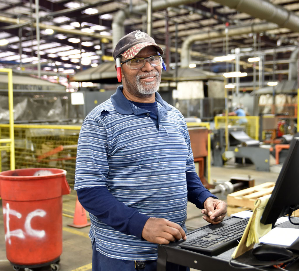 A man stands smiling at a workstation in a factory. Wearing safety glasses, he is dressed in a blue striped shirt and a cap.