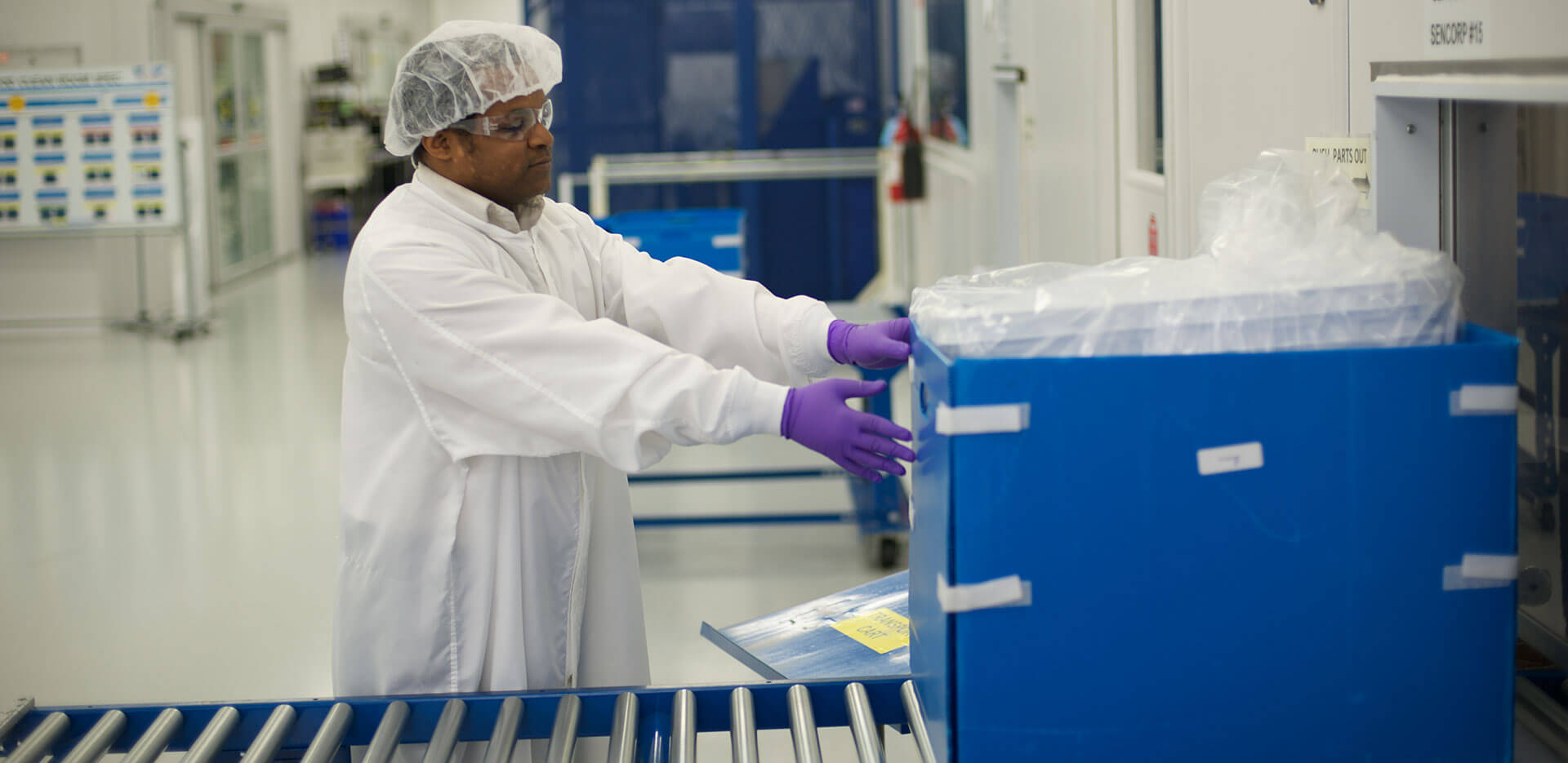 A person wearing a white protective suit, hairnet, and purple gloves works in a clean room. They handle a large blue container wrapped in plastic on a conveyor belt.