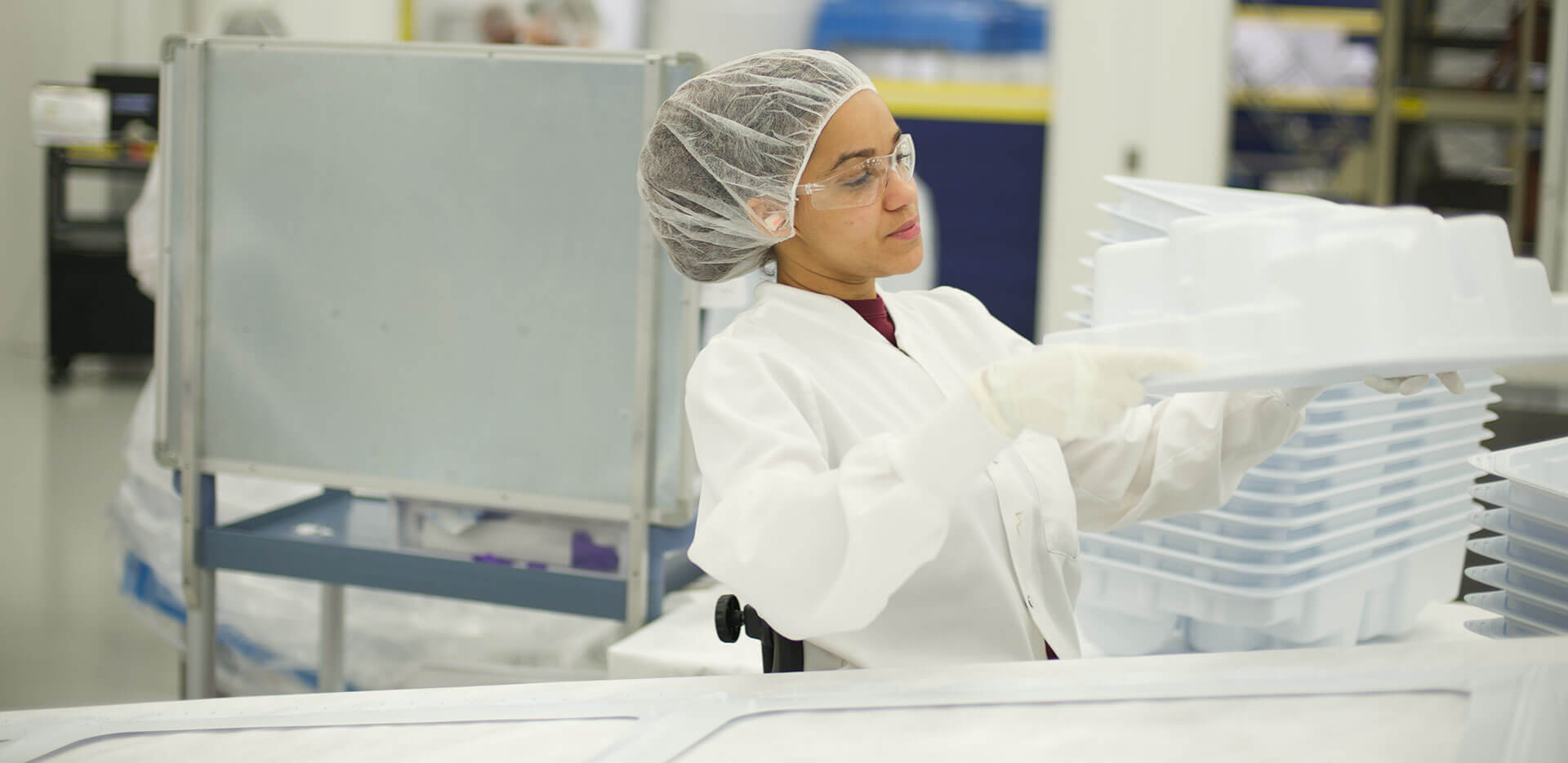 A technician in protective clothing, gloves, and a hair net inspects large plastic trays in a cleanroom environment.