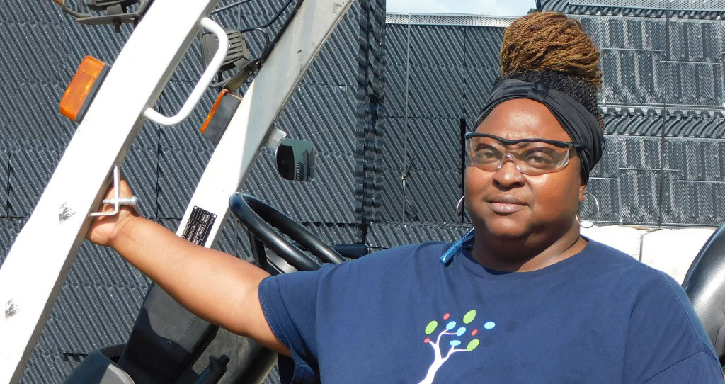 A person wearing safety glasses and a headwrap stands outdoors next to a metal structure, possibly a vehicle. They are dressed in a dark blue shirt with a colorful graphic of a tree on the front.