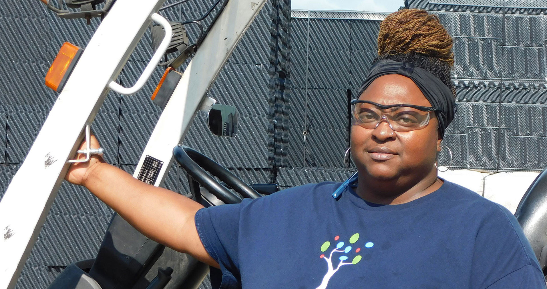 A person wearing safety glasses and a headwrap stands outdoors next to a metal structure, possibly a vehicle. They are dressed in a dark blue shirt with a colorful graphic of a tree on the front.