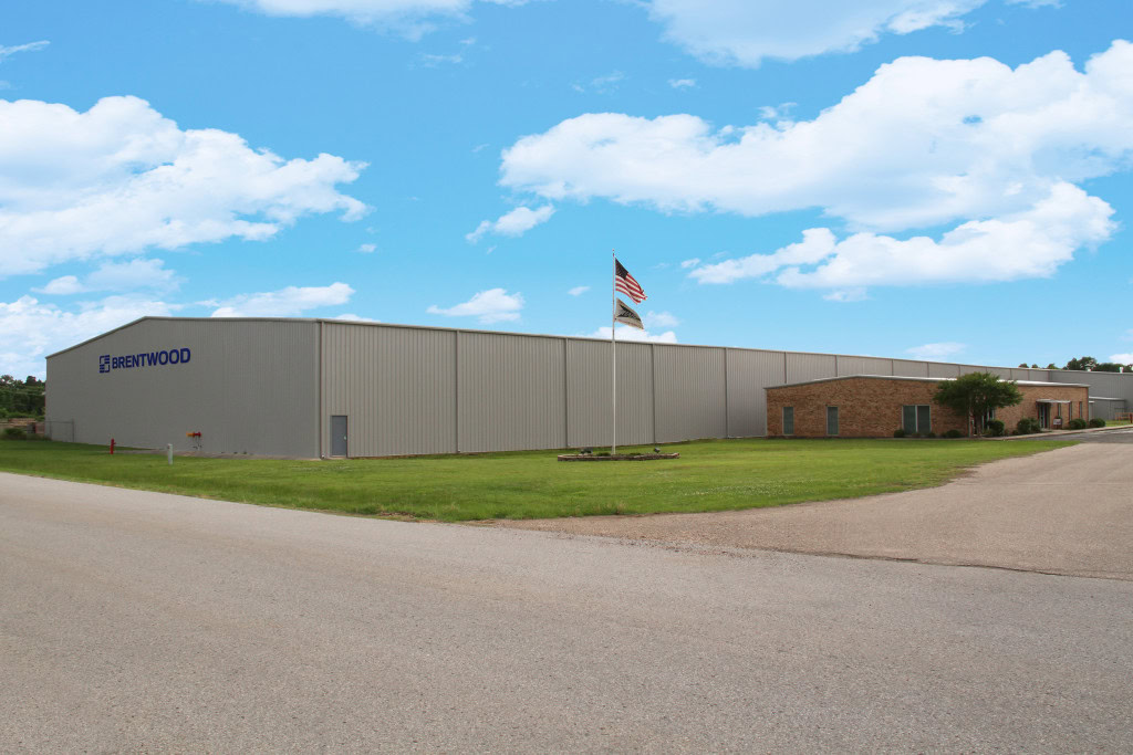 A large industrial building with a "Brentwood" sign on the exterior stands under a bright blue sky with scattered clouds. An American flag is flying on a pole in the front grassy area.