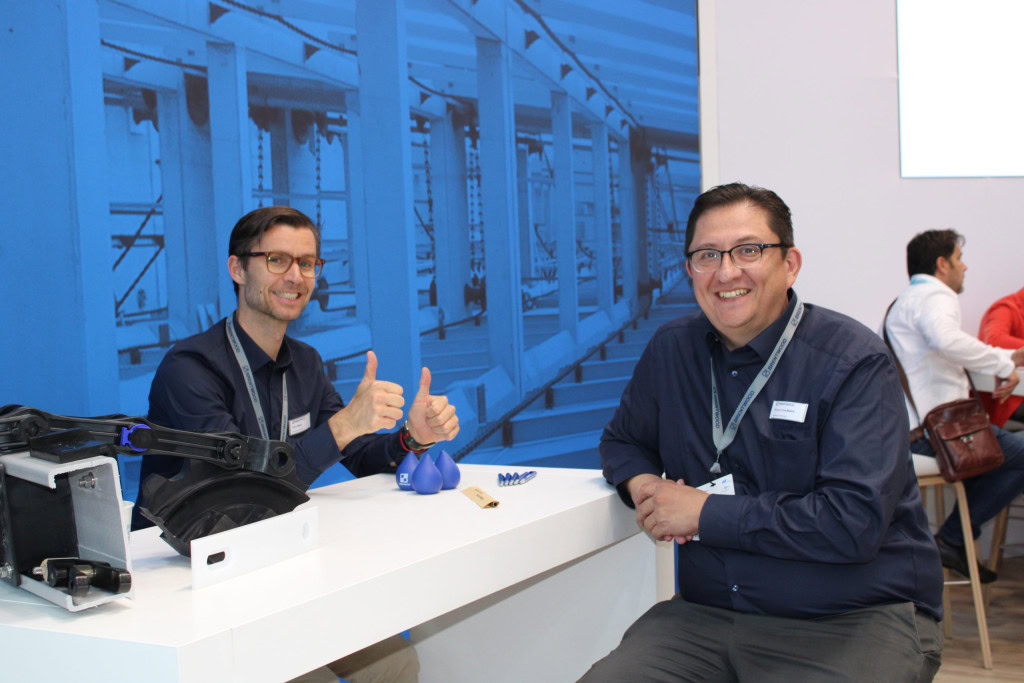 Two men are sitting at a white table in front of a blue-themed backdrop. The man on the left, giving two thumbs up and smiling. The man on the right is also smiling. Both are dressed in dark-colored shirts and wearing lanyards.