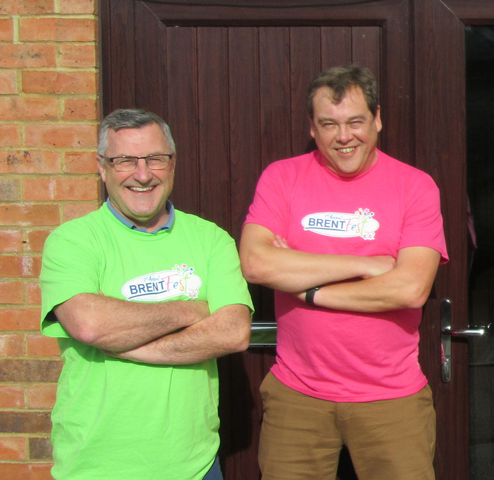 Dos hombres están de pie uno al lado del otro con los brazos cruzados, sonriendo a la cámara. Ambos llevan camisetas de colores -una en verde brillante y la otra en rosa- cada una con el logotipo "BRENTfest".