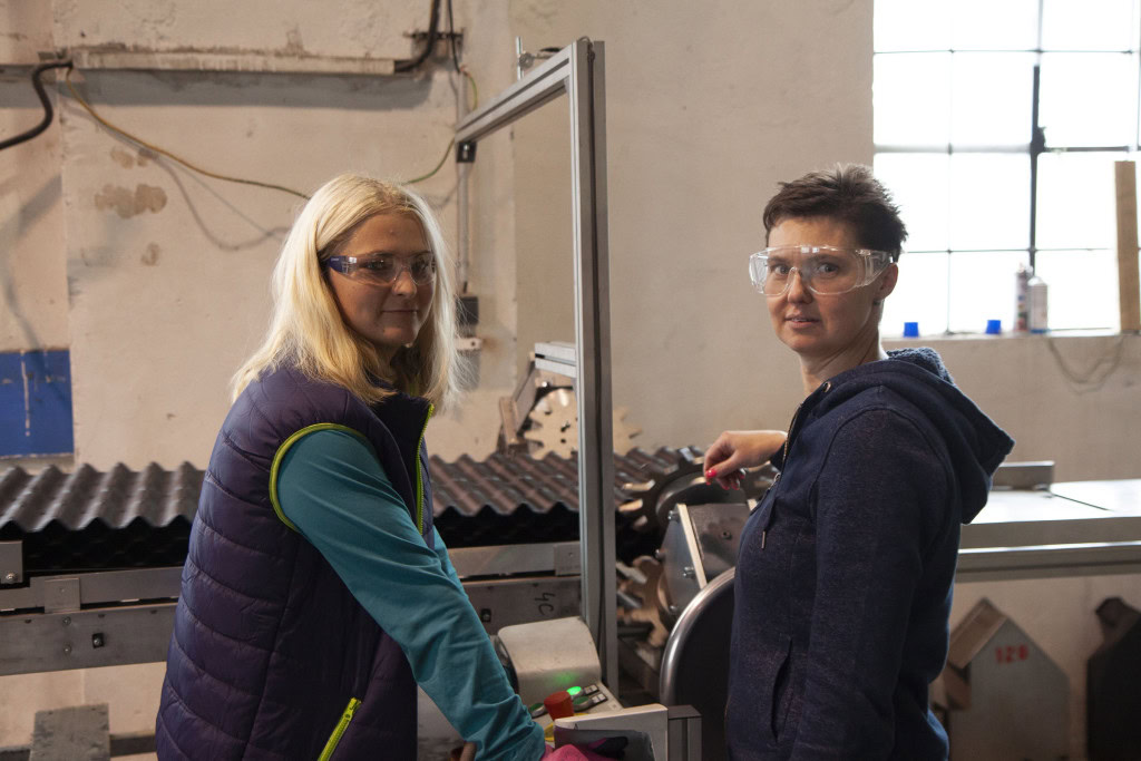 Dos mujeres están de pie una al lado de la otra en un taller, ambas llevan gafas de seguridad. Una tiene el pelo rubio y lleva un chaleco morado, mientras que la otra tiene el pelo corto y oscuro y lleva una sudadera azul con capucha.