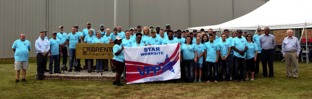 A large group of people wearing matching light blue shirts posing outdoors in front of an industrial building and tent. They are holding up an "Brent Industries" banner and a "Star Worksite VPP" flag.