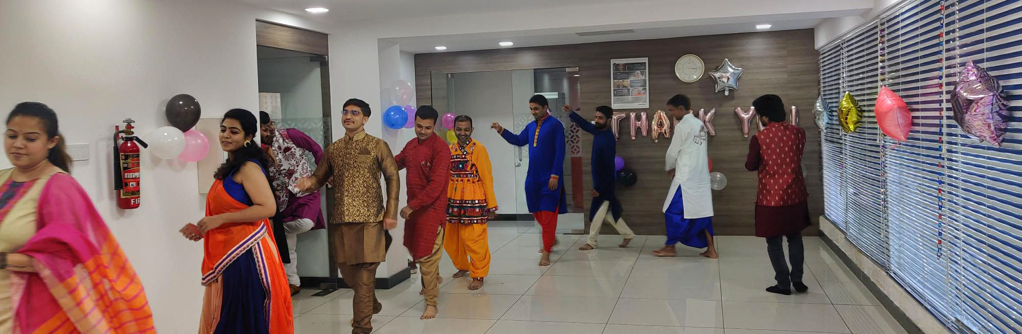 Un grupo de personas vestidas con atuendos tradicionales indios caminan por el interior de una oficina decorada. En las paredes hay globos y una pancarta de "Gracias". Algunos individuos visten colores vibrantes como el naranja, el rosa y el azul.