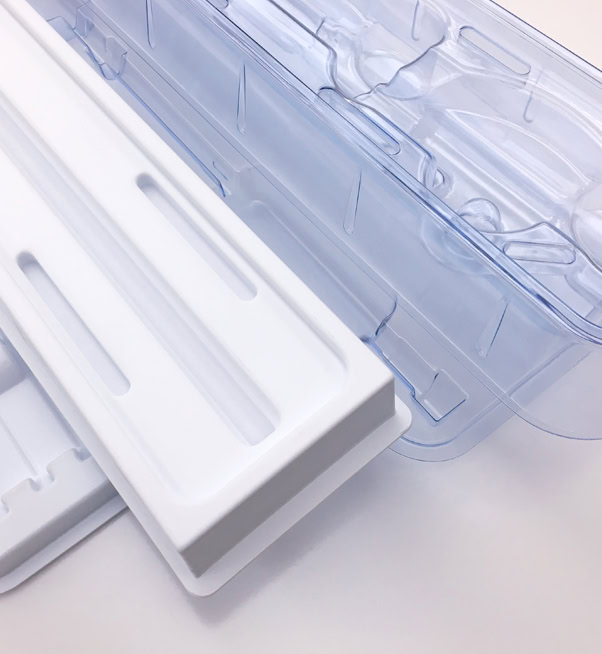 Clear and white plastic trays, are positioned at an angle against a plain white background. These packaging trays have elongated slots and grooves designed for holding items securely.