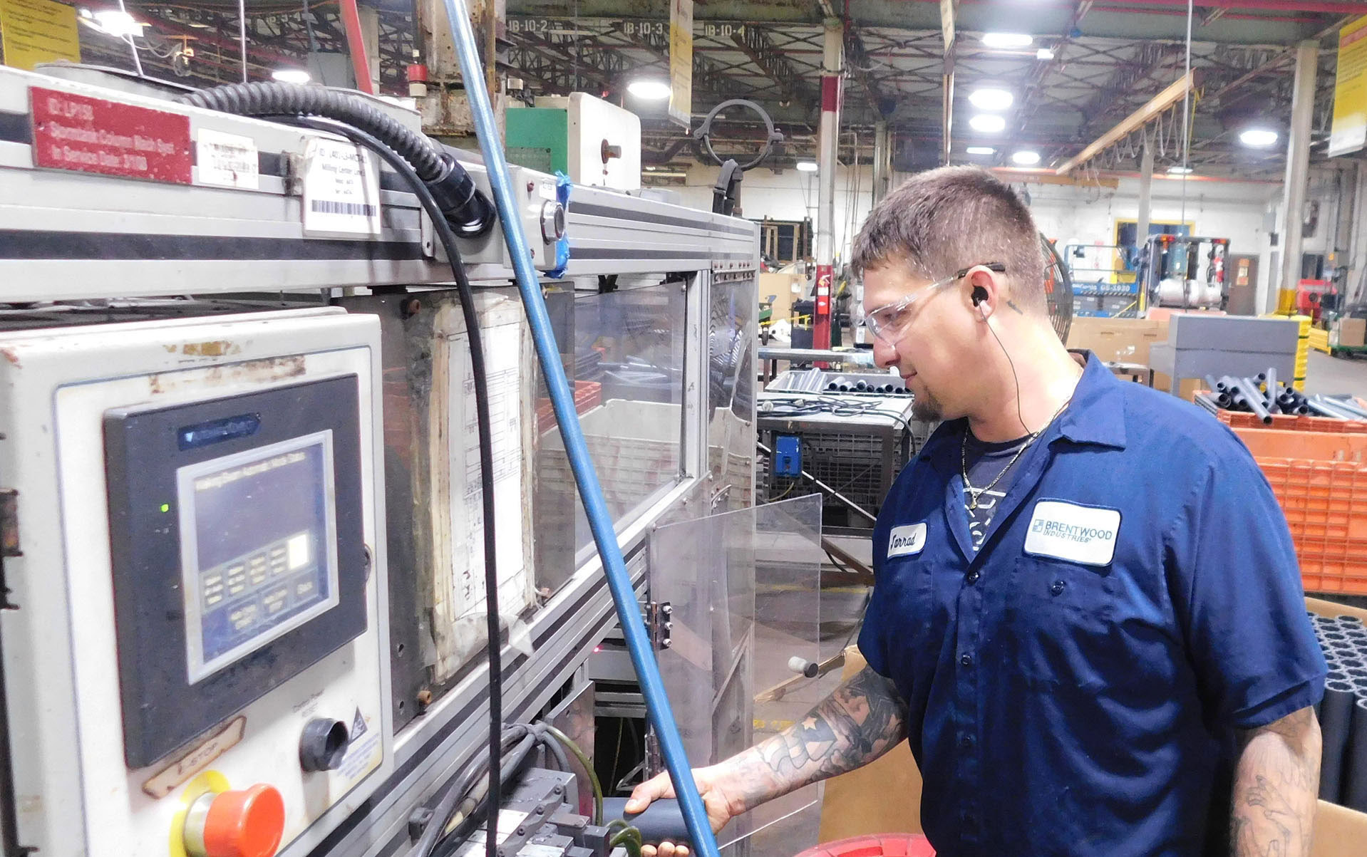 A worker in a blue uniform, wearing safety glasses and ear protection, operates machinery in a factory in Lebanon, Pennsylvania. He adjusts the controls on the machine, while various factory equipment and tools are visible in the background.