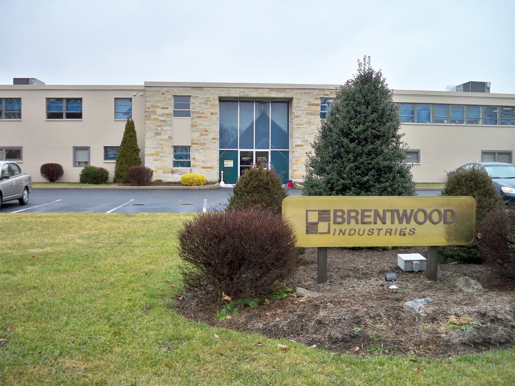 A beige office building with multiple windows and a glass door entrance. In the foreground, a sign reads "Brentwood Industries." The landscaped area features shrubs and small trees, with a few cars parked in front of the building.