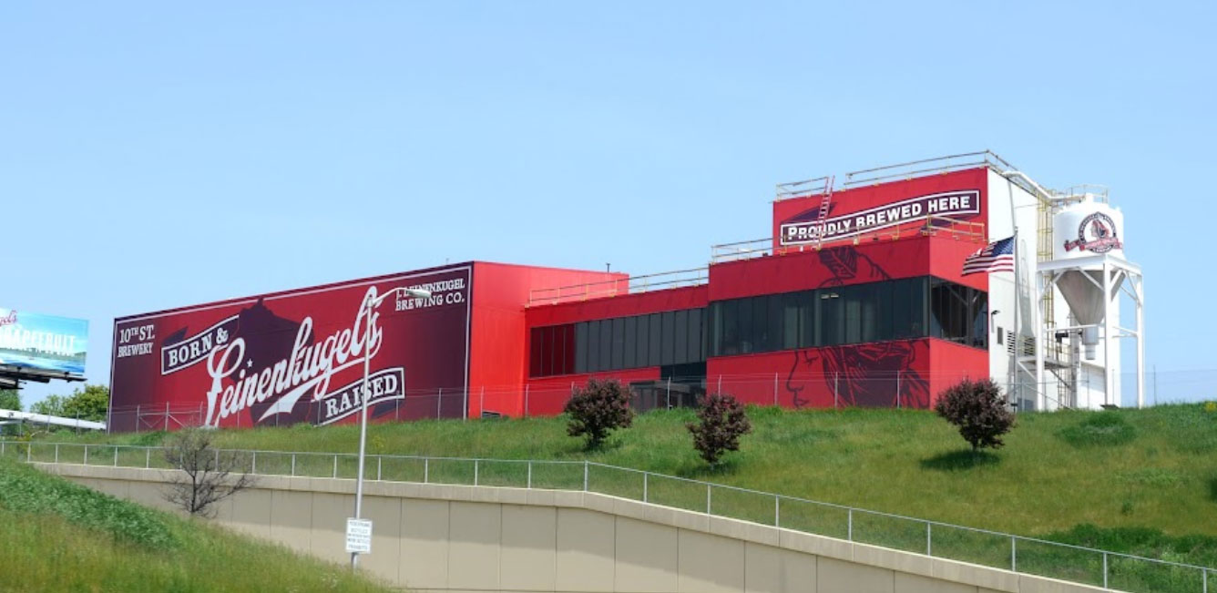 Image of a large, red brewery building for Leinenkugel's in Chippewa Falls, Wisconsin. The well-kept construction showcases the brand's name prominently and a sign reading "Proudly Brewed Here."