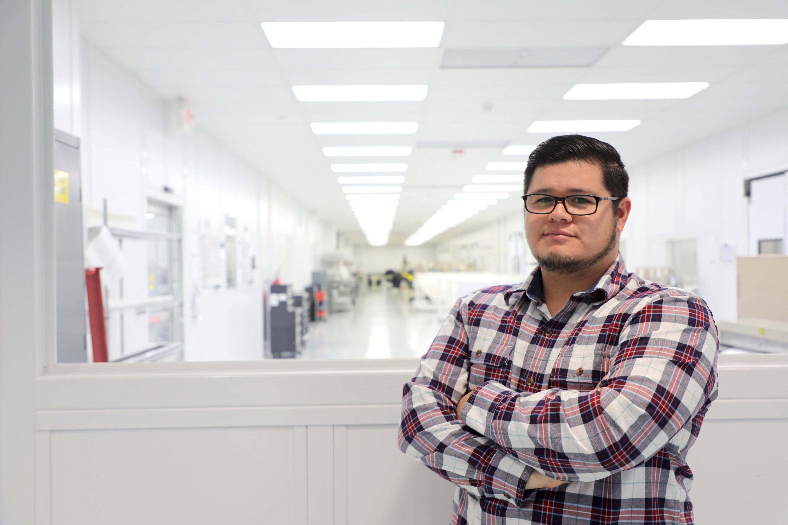 A man with short dark hair, glasses, and a beard stands with his arms crossed, wearing a plaid shirt. He is in front of a large window that looks into a brightly lit, spacious laboratory in Tijuana, Mexico, featuring white walls and ceiling lights.