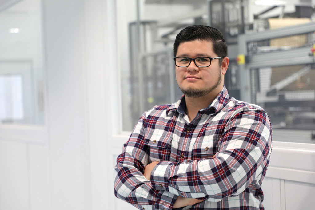 A man with short dark hair, glasses, and a trimmed beard, wearing a plaid shirt, stands with his arms crossed in a modern office or industrial space in Tijuana, Mexico, with machinery visible through the windows in the background.