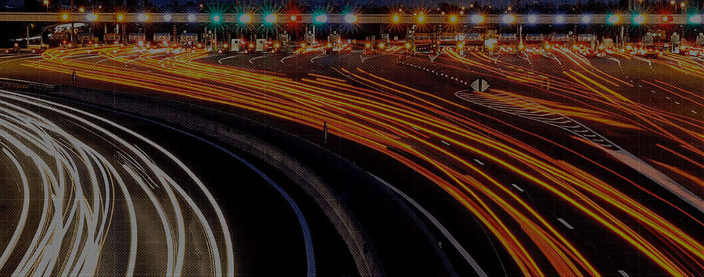 Long exposure photo of toll booths at night, showing the continuous streams of car headlights and taillights forming curving light trails.