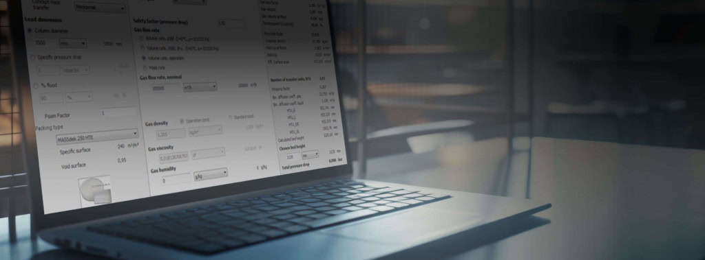 A laptop sits on a dark, wooden table in a dimly lit room. The laptop screen displays a software interface with various numerical data and dropdown menus, including calculations for mass transfer.