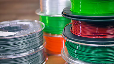 Close-up of multiple spools of filament in various colors, including gray, orange, green, and red. The spools are stacked and arranged randomly on a table.