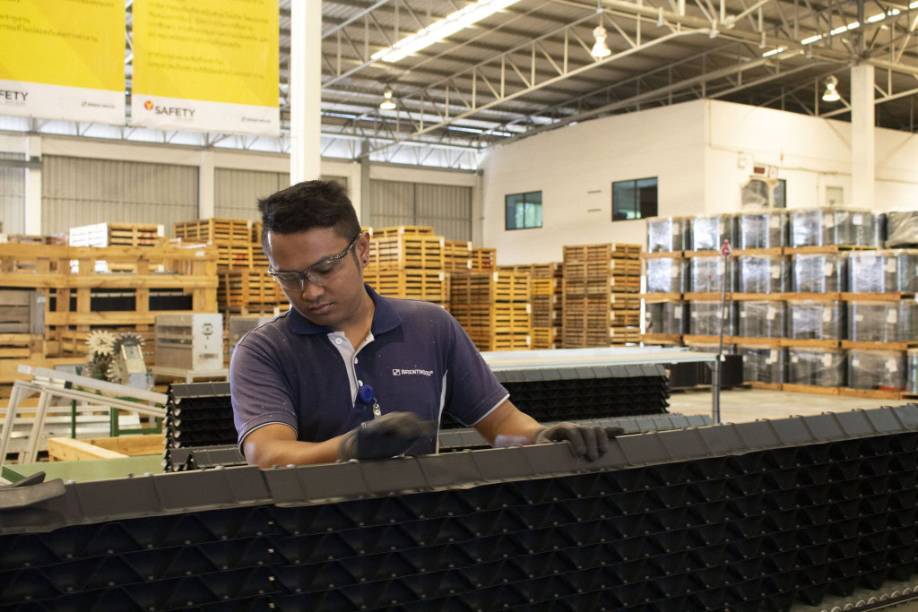 A person wearing safety glasses and gloves works on an assembly line in a warehouse. Around the area, stacks of wooden pallets and wrapped goods are visible.