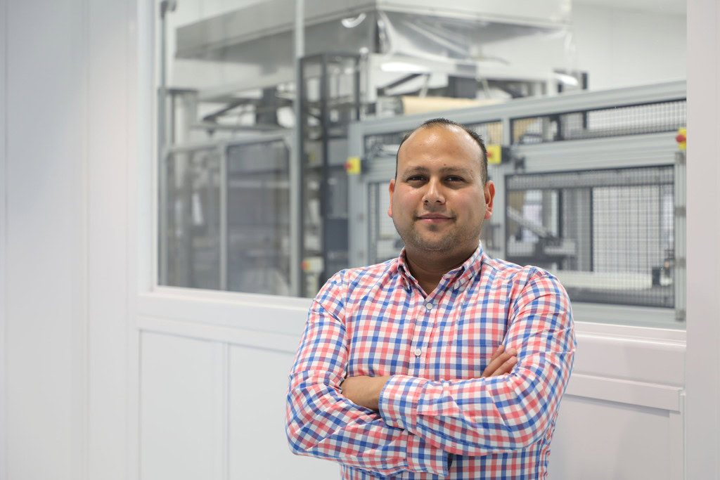 A man in a plaid shirt stands confidently with arms crossed in front of a modern industrial machine behind a glass partition.