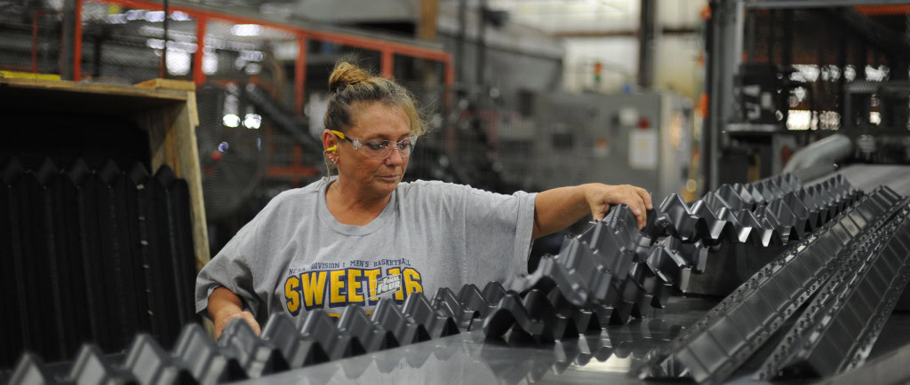 A worker in a gray shirt and safety glasses inspects black industrial parts in a factory setting, with machinery in the background.