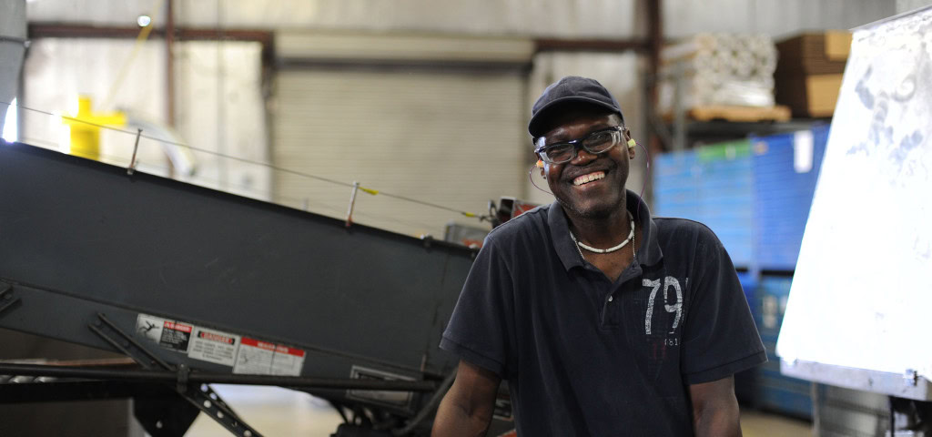 A man wearing a black cap and dark polo shirt smiles while standing in an industrial setting. Behind him is various machinery and equipment.