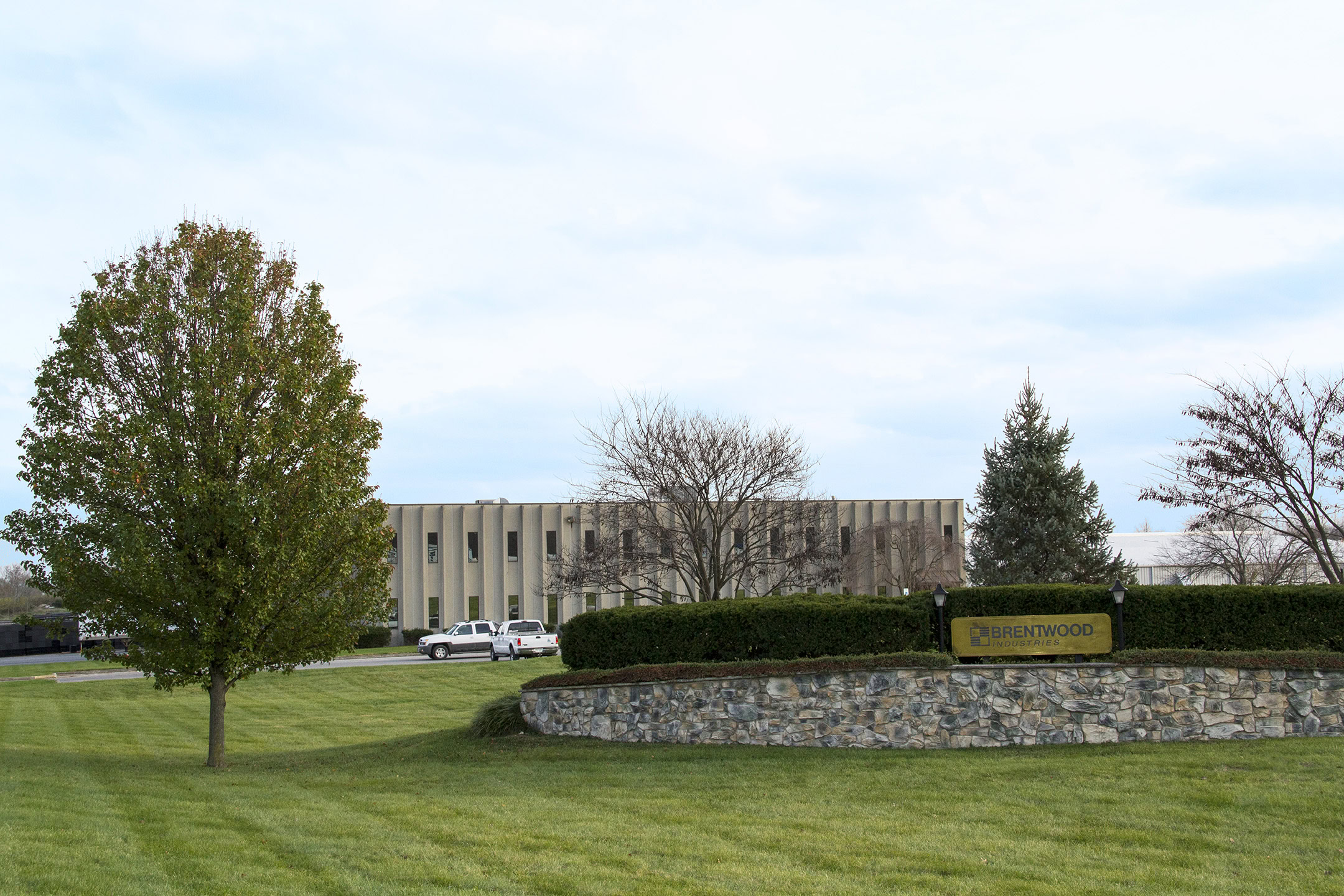 A large office building is in the background with a beige facade. In the foreground, there is a stone sign that says "Brentwood" surrounded by neatly trimmed hedges.