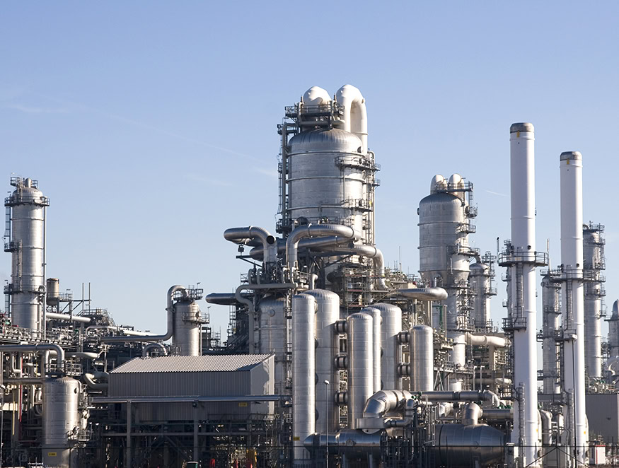 An industrial complex featuring multiple large, cylindrical metal structures, pipes, and chimneys set against a clear blue sky.