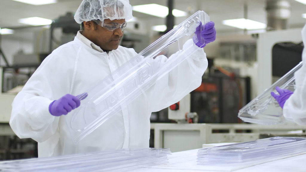 A worker in a cleanroom environment, wearing a white lab coat, hair net, safety goggles, and purple gloves, inspects a large, transparent plastic component at a manufacturing facility.