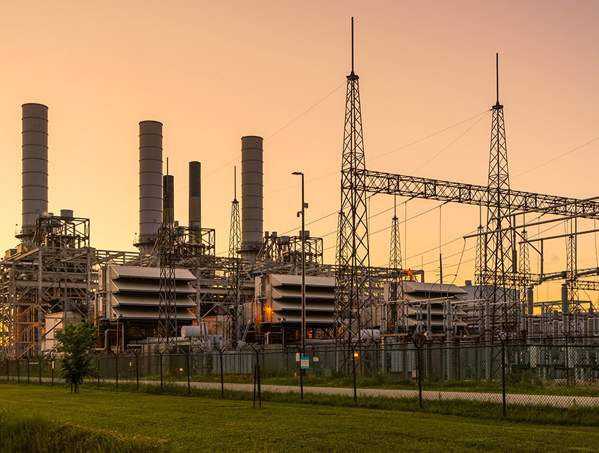 A power plant with multiple industrial structures and tall chimneys stands behind a chain-link fence. Electrical towers and wires extend across the facility.