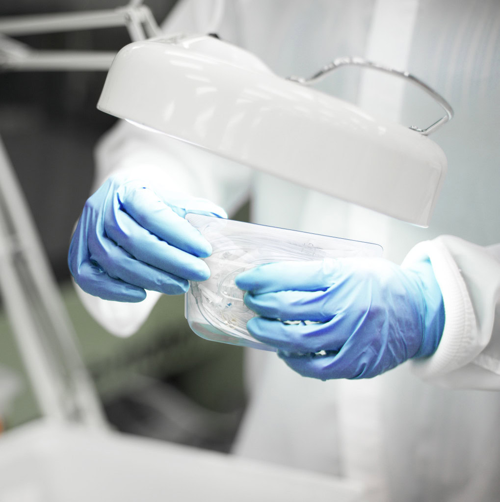 A person in a lab coat and blue gloves examines a transparent container under a bright lamp in a medical laboratory setting. The container appears to hold some form of specimen or sample related to healthcare research.