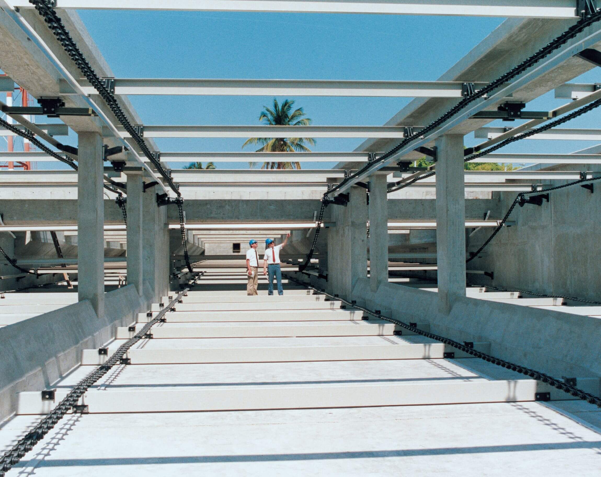 Two workers with helmets stand in the middle of a large construction site framed by concrete beams and structures.