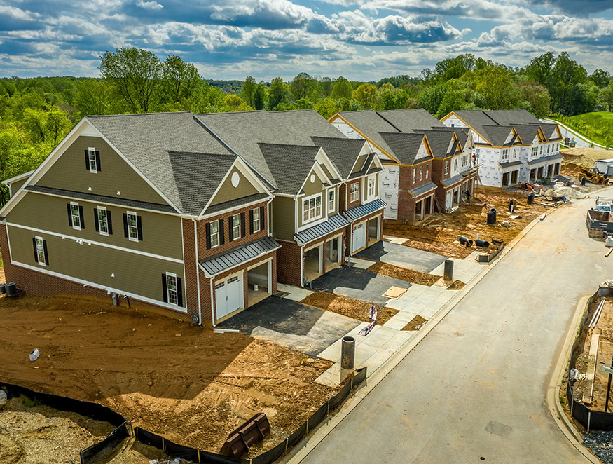 This aerial image shows a row of townhouses under construction in a suburban neighborhood. The left side features nearly completed homes with landscaping and driveways, while the right side has houses in various stages of construction.