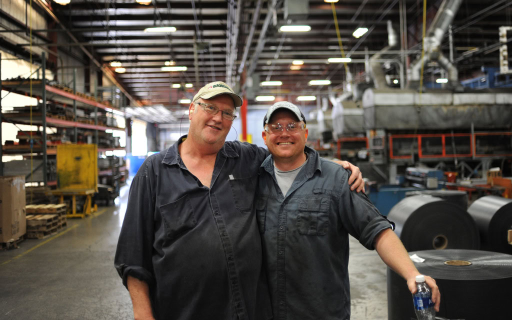 Two factory workers wearing hats and safety glasses stand inside an industrial facility. They are smiling with their arms around each other's shoulders.