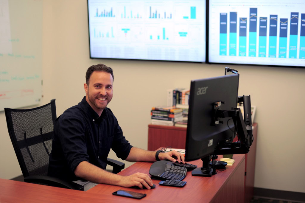 A smiling man sits at a desk, working on a computer with a dual-monitor setup. Behind him, two large screens display various data charts and graphs.