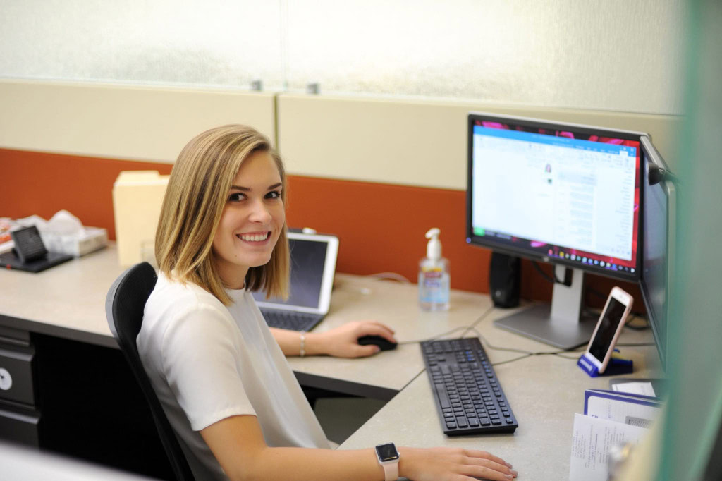 A woman with shoulder-length blonde hair sits at a desk in an office cubicle, smiling at the camera. She works on a computer with a monitor, keyboard, and mouse.