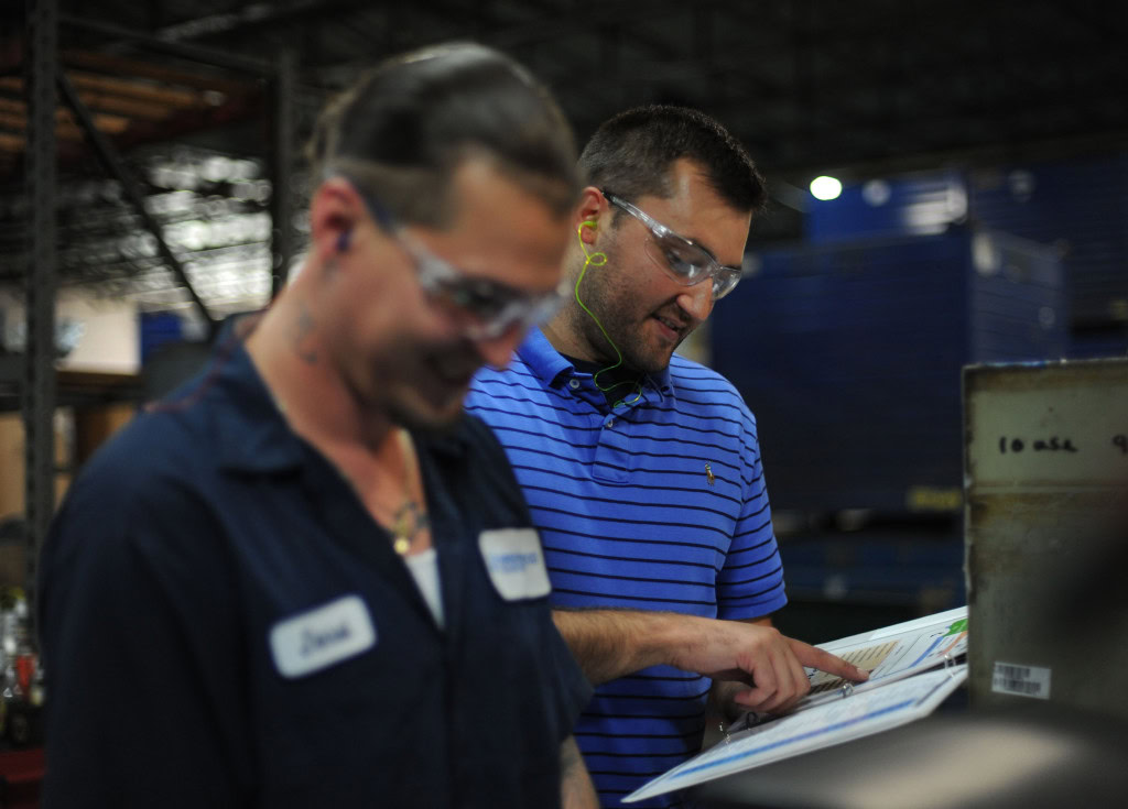 Two men are working in an industrial setting. The man on the left, sporting dark work clothes, discusses a document with the man on the right, who is donning a blue polo shirt and safety glasses.