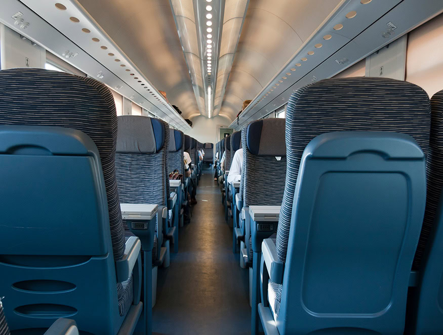 The interior of a train cabin is shown with rows of blue seats on either side of a central aisle. The seats have built-in screens on their backs.