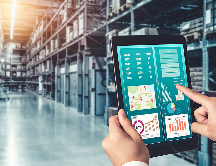 Una persona sostiene una tableta que muestra datos logísticos, incluidos mapas, diagramas de barras y gráficos, mientras permanece de pie en un almacén grande y organizado con estanterías repletas de diversos artículos.