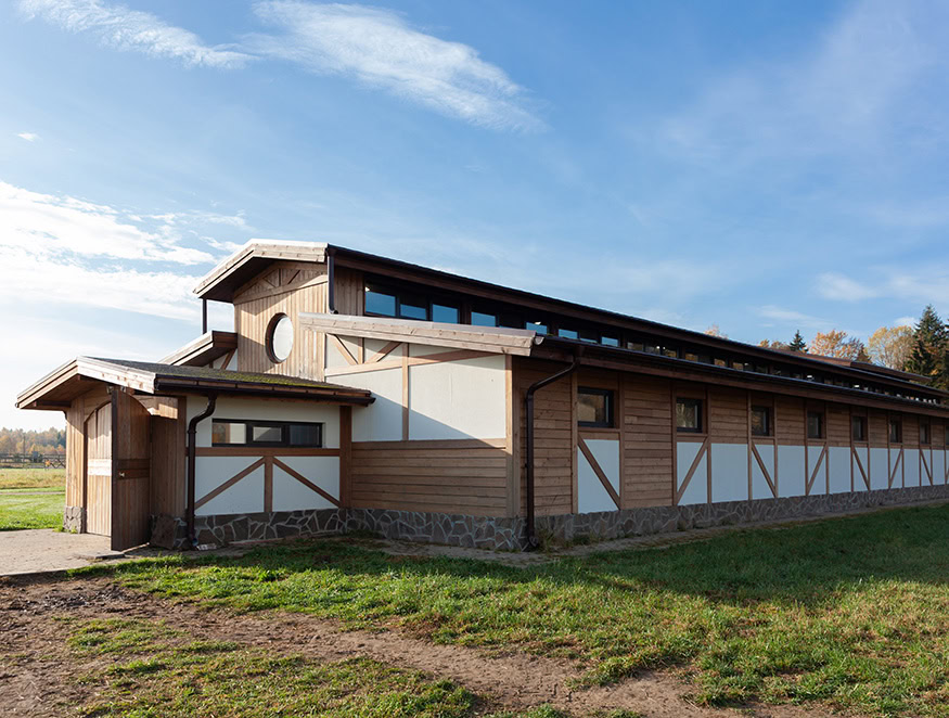 A single-story, modern barn with a mix of wood and white paneling stands under a clear blue sky. The structure features multiple small windows along its length and a circular window near the roof.