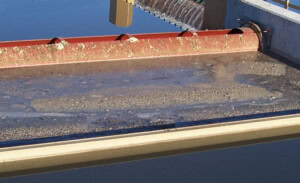 A close-up of water flowing over a weir at a wastewater treatment plant. The image shows sediment and debris, part of the scum removal process, on the water surface as it moves over a red and brown barrier before falling into a lower section. The structure is surrounded by concrete.